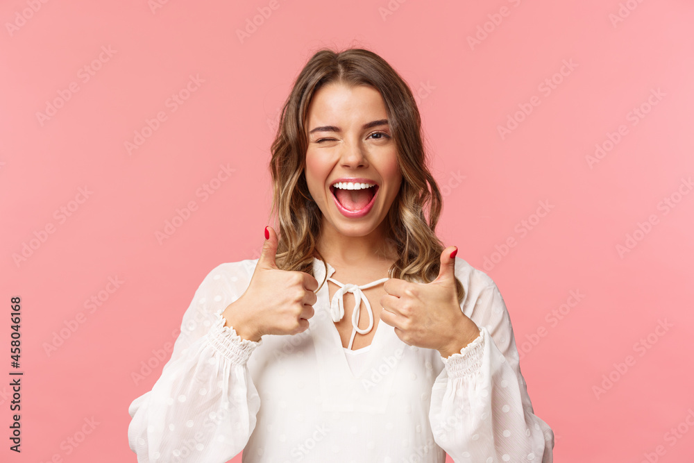 Portrait of sassy good-looking blond girl assure all good, show thumbs-up and wink with classy pleased smile, recommend visit place, shop at online store, standing in white dress pink background